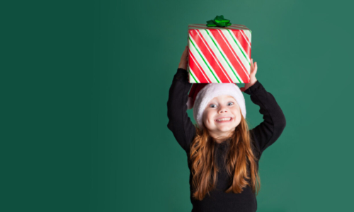 Color image of an excited 4-year-old girl wearing a Santa hat and holding a Christmas present on a green background.
