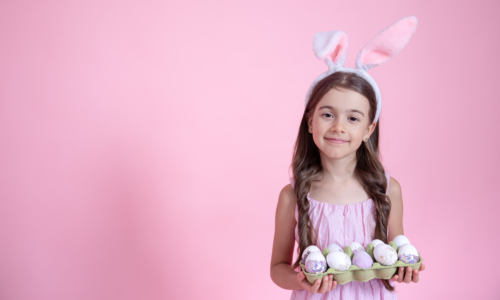 Cheerful little girl with Easter bunny ears and a tray of eggs in her hands on a pink studio background. Easter holiday concept.